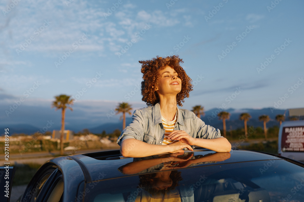 Fototapeta premium Young woman leans on a car roof, smiling with eyes closed in a sunlit outdoor moment, palm trees in the distance, warm breeze and relaxed, casual vibe
