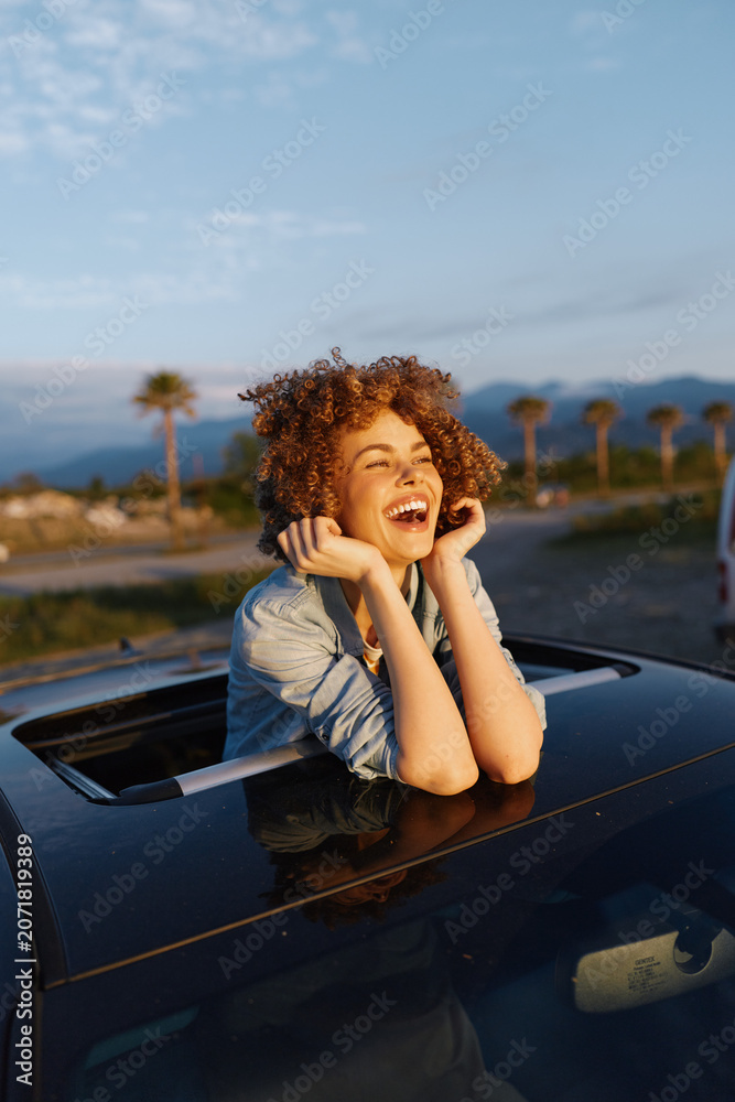 Fototapeta premium A joyful woman leans from a convertible, smiling with hands near her face, enjoying golden hour light. Outdoor lifestyle shot conveying happiness, vacation, travel, and freedom.