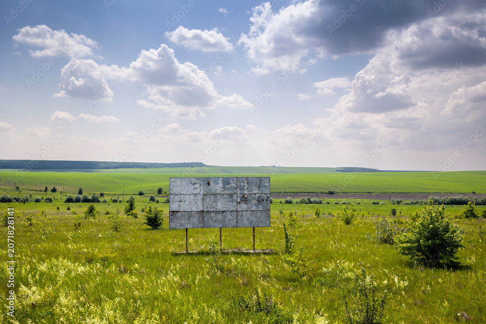 Obraz premium A weathered, blank billboard stands in a vast grassy field with scattered bushes under a partly cloudy sky. Expansive green agricultural fields stretch towards a distant treeline at the horizon
