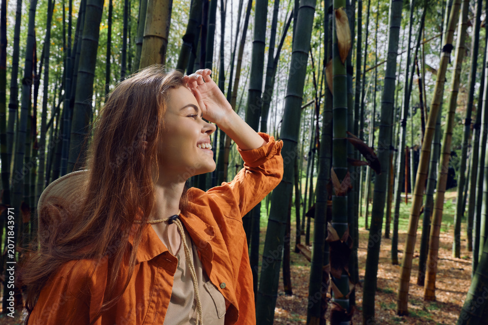 Fototapeta premium Woman portrait in a bamboo forest with a bright smile, wearing a straw hat and orange jacket, sunlight filtering through stalks, nature scene conveying joy and peaceful outdoor exploration.