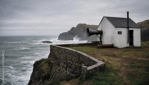 A small white stone building with a large metal foghorn on a cliff edge overlooking a stormy ocean with crashing waves and rocky coastline