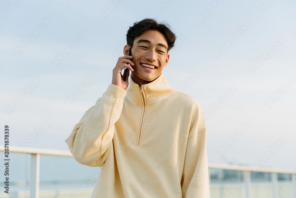 Obraz premium Portrait of a cheerful young man on a balcony speaking on phone in a soft yellow pullover for lifestyle background and banner use