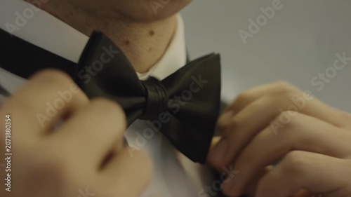 Male adjusting black bow tie on white dress shirt, focusing on hands and neck area, preparing for formal event in a softly lit indoor setting