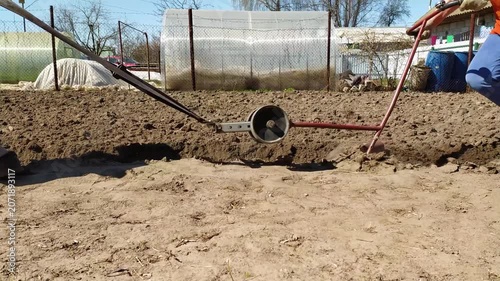 Two farmers work together using manual agricultural tools to till the soil in a garden plot on a sunny day.