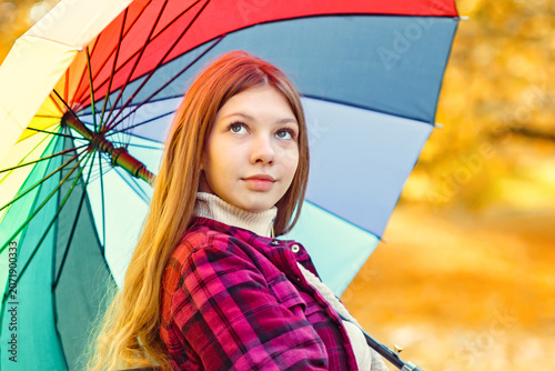 Young person sits under a colorful umbrella looking up at the trees in a park during autumn sunlight
