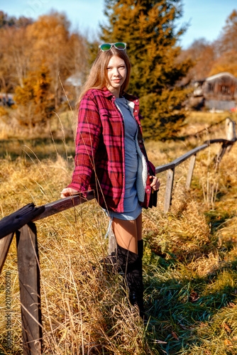 Young woman stands beside wooden fence in grassy field during sunny day in autumn season