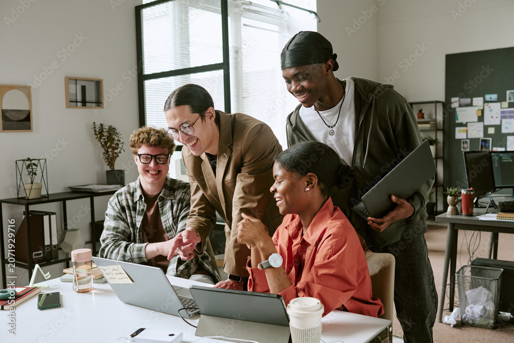 Fototapeta premium Diverse young adult team collaborating around laptop, reviewing project ideas and sharing feedback in office. Ideal for workplace teamwork, startup culture, innovation marketing
