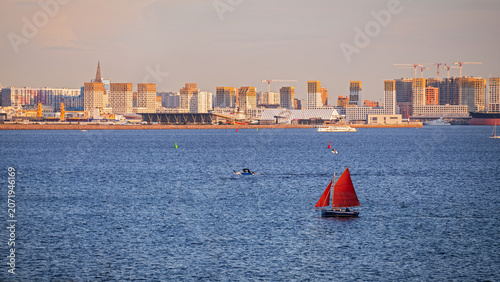 Small sailboat with red sail floating on sea water near Saint Petersburg coastal city skyline.
