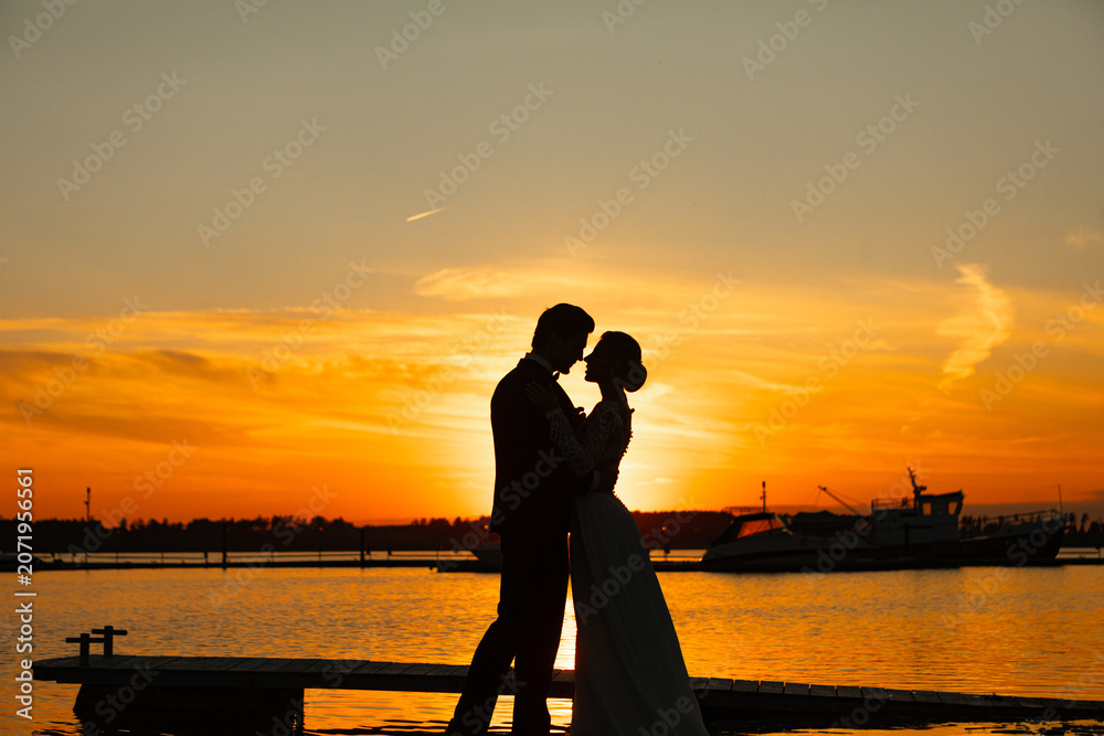 Fototapeta premium Wedding couple silhouette embracing at sunset on marina pier