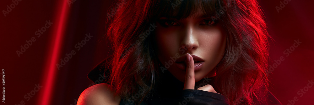 Naklejka premium secretive woman in red glow portrait, enigmatic female making hush gesture, mysterious model in cinematic red light