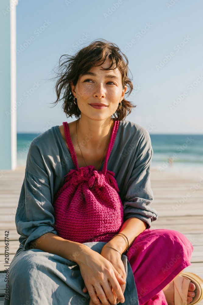 Fototapeta premium Seated woman portrait with a magenta knit bag and relaxed grey outfit on a sunlit pier for lifestyle banner and fashion editorial use