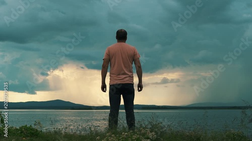 A lone man stands on the shore of a lake against a cloudy, stormy sky. Human psychology, time alone with oneself.