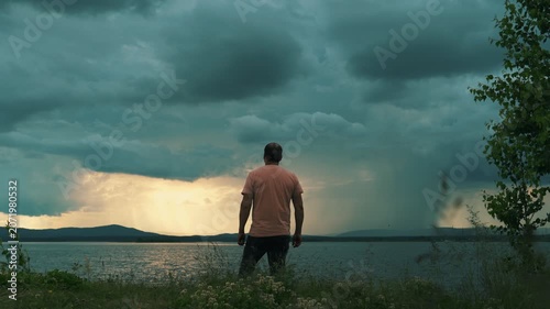 A lone man stands on the shore of a lake against a cloudy, stormy sky. Human psychology, time alone with oneself.