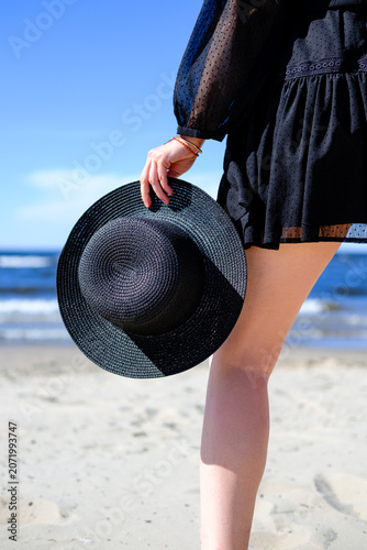 Close-up of a woman in a black dress holding a black straw hat while standing on a sandy beach. Summer vacation concept with blue sky, sea waves, and elegant fashion style.
