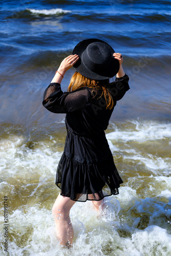 Rear view of a woman wearing a black dress and black hat while standing in ocean waves. Summer vacation and freedom concept with dynamic water splashes and seaside atmosphere.