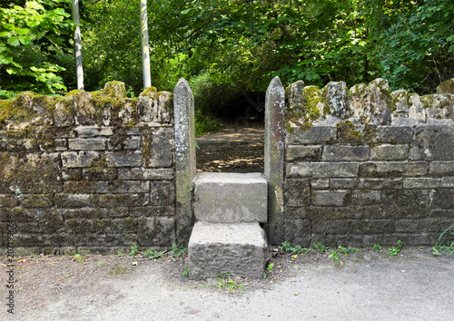A traditional stone stile cuts through an old dry stone wall, opening into woodland with a quiet lake, lush greenery, and wild plants in Horton Bank Country Park, Clayton, Yorkshire, UK