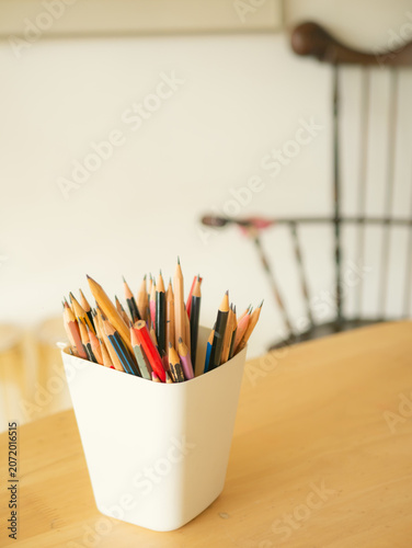 Colorful Pencils in White Cup on Wooden Table with Blurred Chair Background Copyspace