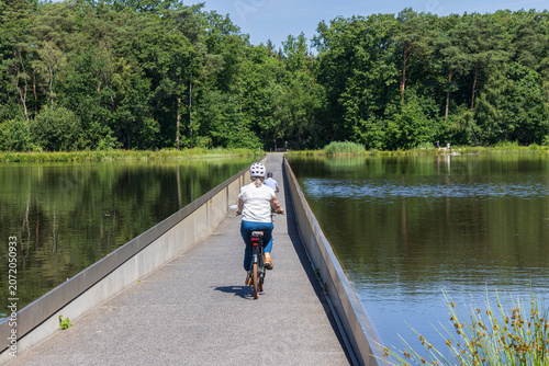 Couple cycling across narrow causeway through a lake in Bokrijk park near Genk, Flanders in Belgium