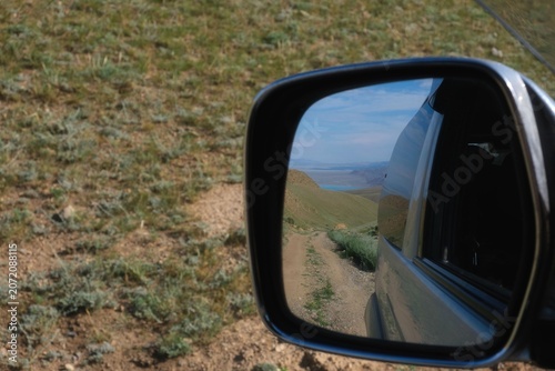 Rear view car mirror reflecting a dirt road leading to a distant lake in a hilly landscape. Commercial travel background with copy space, ideal for road trip and adventure lifestyle concepts.