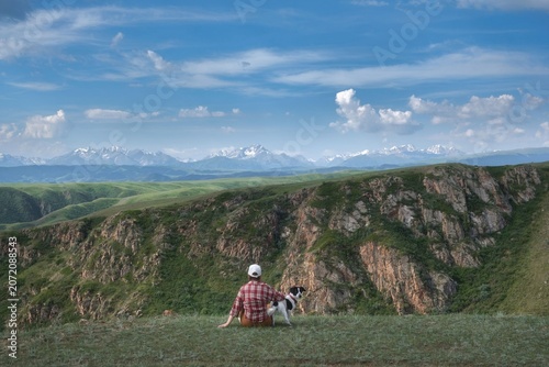Woman with dog sits on green grass cliff looking at distant snow mountain range. Commercial travel lifestyle background with copy space for outdoor adventure concept.