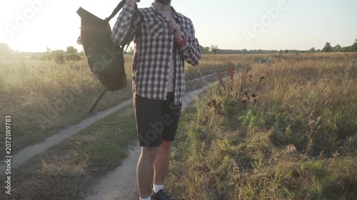 A cyclist in a helmet rolls down and closes his black roll-top backpack, puts it on, and walks out of the frame to continue his bicycle journey along a field trail.