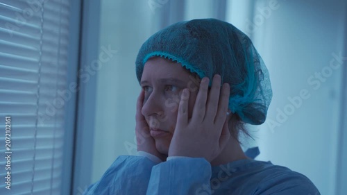 A traumatized woman with a black eye and facial bruises stands in a hospital corridor wearing a medical gown and cap, looking shocked and deeply suffering after domestic violence.