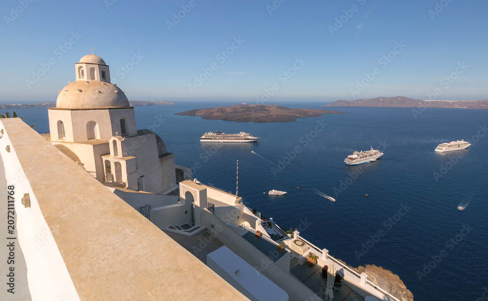 Obraz premium Whitewashed church dome overlooking the Santorini caldera with cruise ships anchored in the Aegean Sea.