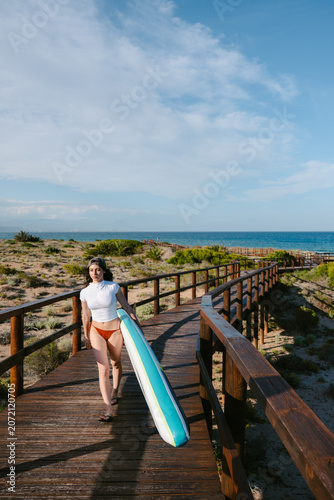 Woman carrying paddleboard walking wooden beach walkway