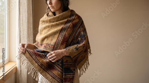 Woman wearing a woven textile shawl with autumn season patterns standing near a window for a cozy interior lifestyle concept