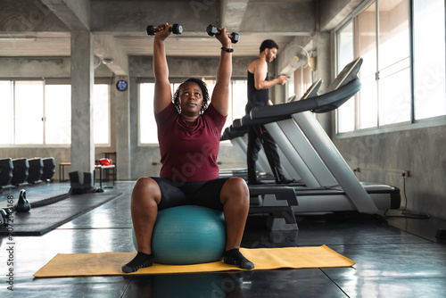 Obraz na płótnie Focused curvy African American woman lifting dumbbells overhead while sitting on a blue stability ball in a modern industrial gym, demonstrating strength, health, and inclusive fitness motivation