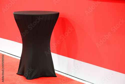 Black fitted cocktail table with stretch draped cloth standing against a bright red wall and white baseboard on red carpet