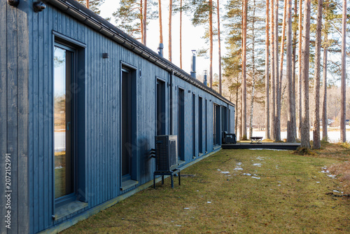 Slika na platnu A long single story cabin with dark cladding and tall windows extends beside straight pine trees, lingering snow, exterior heat pumps, and stacked patio chairs in cool light
