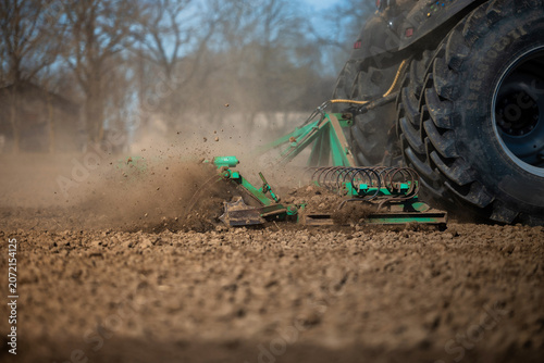 Εκτύπωση καμβά Ground level view shows a tractor pulling green tillage implements as dust rises, soil clods break, and coil springs and treaded tire stand out in daytime light