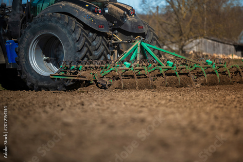 A large tractor tows a green spring tooth harrow across a brown field, dust flies, metal tines rake soil, a weathered barn and leafless trees stand under daylight.