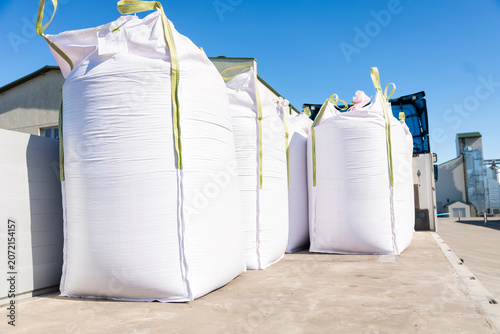 Oversized white FIBCs with pale green loops stand on a concrete dock near a silo and forklift mast at an industrial facility in daylight, viewed from a low angle.