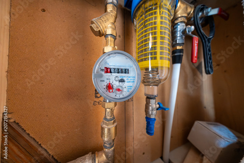 Close up shows an analog water meter with m3 marking, brass fittings, and a transparent sediment filter housing with yellow cage and blue drain valve indoors in natural light.