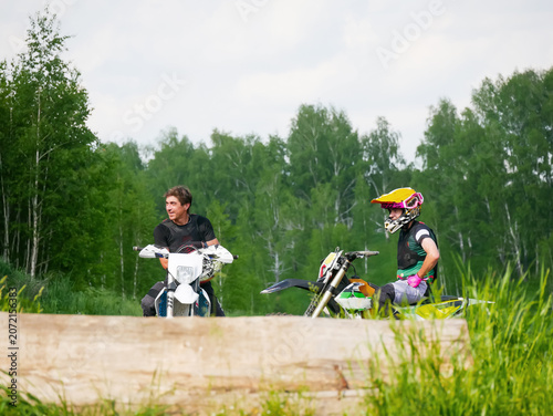 Two motocyclists sitting on dirt bikes in forest. Man and teenager wearing protective gear before motocross training. Extreme outdoor sport, hobby, adventure and lifestyle in nature.