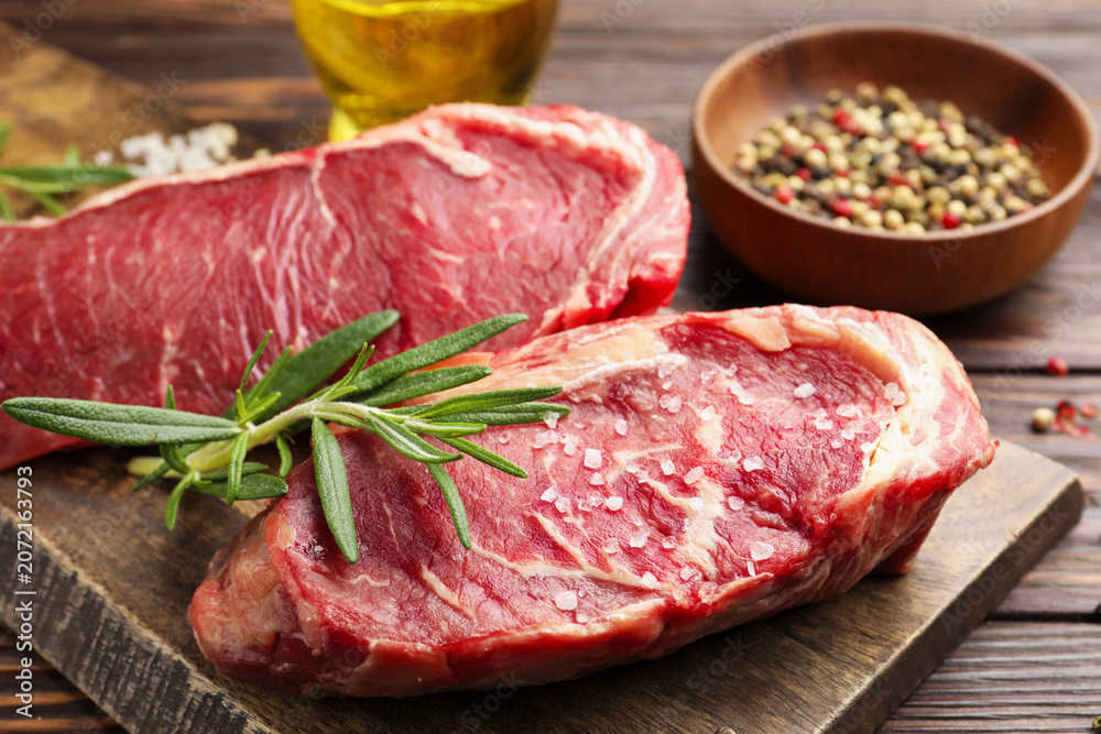 Fototapeta premium Raw beef steaks, spices, rosemary and oil on wooden table, closeup