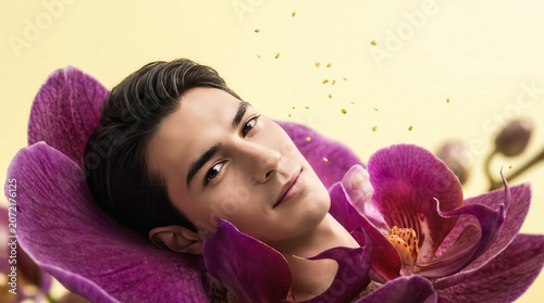 Soft light bathes a serene, calm young man. He lies nestled within vibrant purple orchids. His gaze is gentle, his expression peaceful and trusting. Petals surround him. Ideal for wellness, beauty