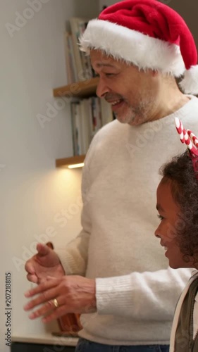 Vertical video: Clapping senior in holiday headwear offering cookie in kitchen, child leaning