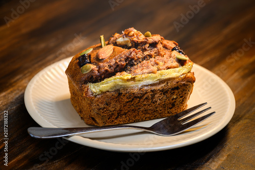 Close up of homemade banana bread topped with mixed nuts and seeds, served on a plate with a fork on a wooden table. Delicious healthy dessert concept.