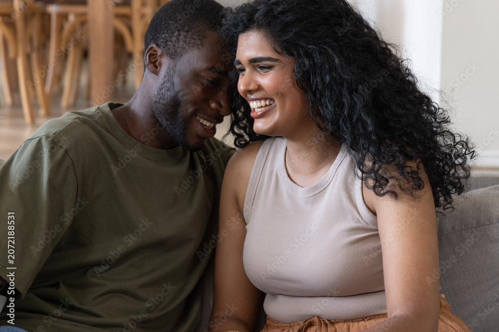Obraz premium Diverse couple laughing and leaning together on light sofa in living room, wooden table behind