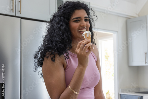 Adult Indian woman holding soft-serve cone, smiling in tank top, gold bracelets in home kitchen