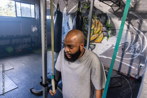 Fotografie Man with beard wearing grey t-shirt standing near squat rack with suspension str