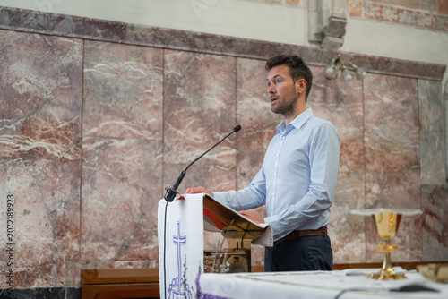 Schilderij op canvas Preacher standing behind a pulpit with a microphone in a church, reading from a Bible to deliver a sermon, homily, or lead prayer