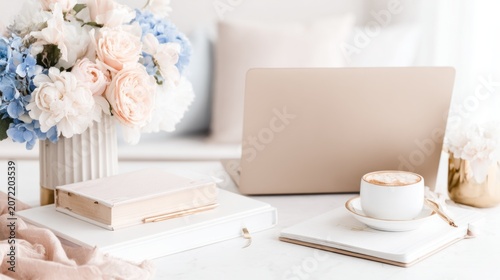 A clean desk displays a laptop, warm cappuccino, colorful flowers, a book, and a notebook