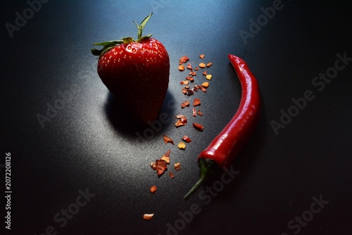 Artistic dark food composition featuring fresh strawberry, red chili pepper and chili flakes on black background.