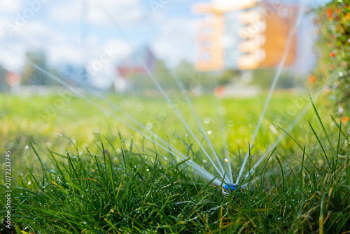 Automatic irrigation system sprays water across grassy lawn in outdoor garden environment. Fresh grass water droplets and irrigation hardware operating in residential or public green space. Natural