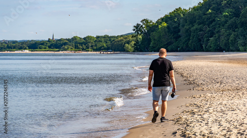 Man Walking Along Baltic Sea Beach Carrying Headphones on Sunny Summer Day