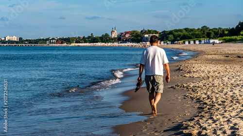 Man walking barefoot along Baltic Sea shoreline carrying sandals